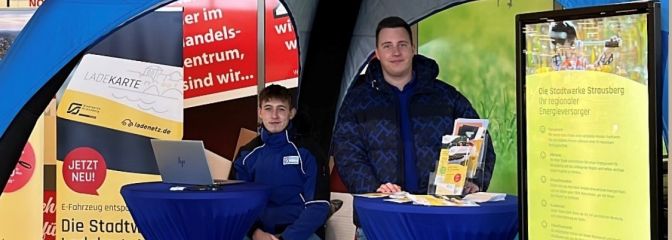 2 Männer sitzen an blauem Infostand an einem Tisch und lächeln