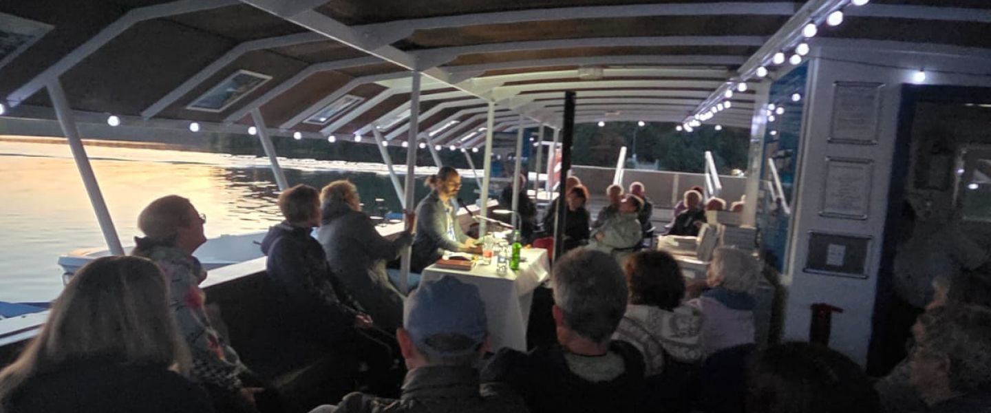 abendliche Lesung, beluchteter Tisch mit Menschen auf Stühlen von hinten